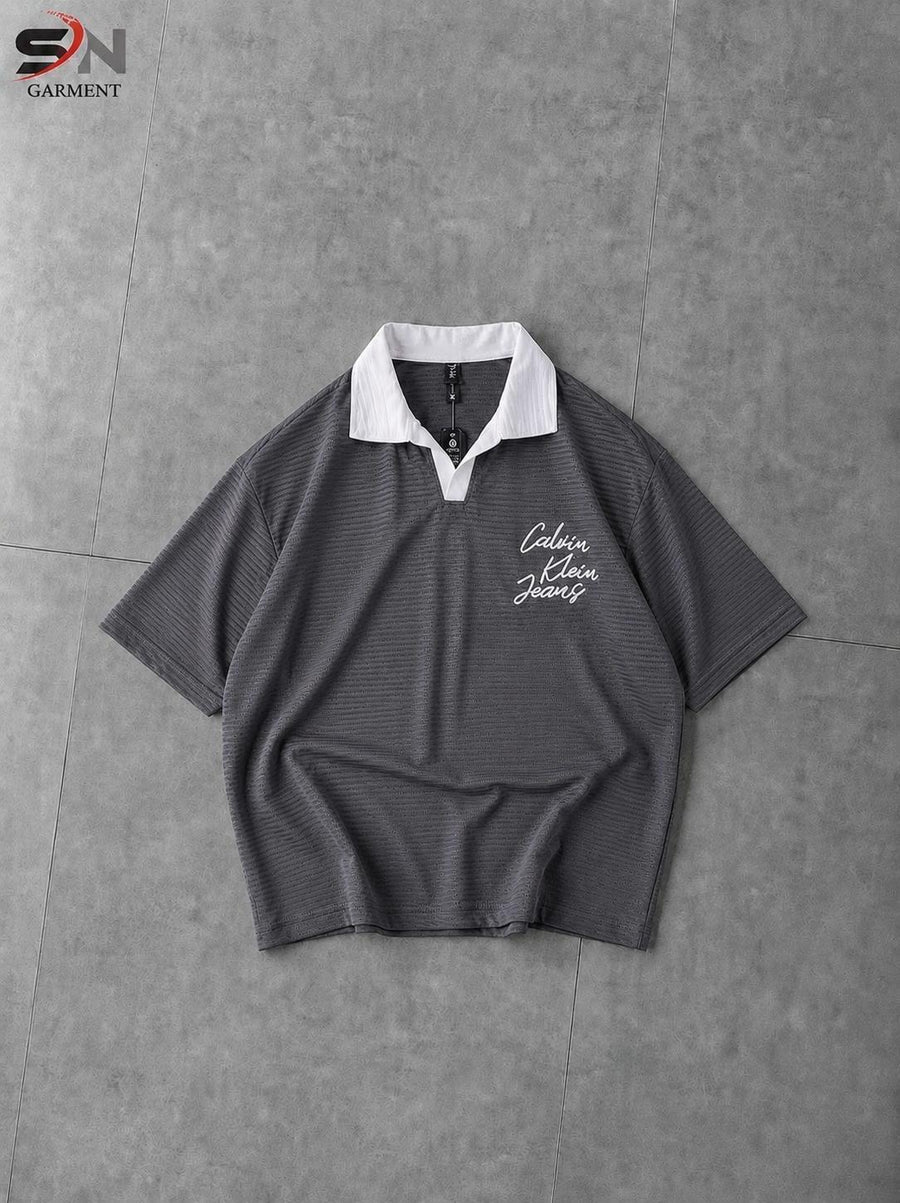 A dark gray short-sleeve polo shirt with a white collar and white script embroidery on the left chest, laid flat on a gray tiled surface.