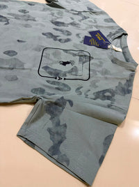 A light gray short-sleeve t-shirt with a dark gray camouflage pattern, featuring a black graphic print of a polo player inside a rectangular frame on the chest. A blue Polo Ralph Lauren brand hangtag is attached near the collar, and the shirt is laid flat on a light beige tiled floor.