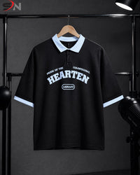 A black short-sleeve polo shirt with a light blue collar and sleeve cuffs, featuring white graphic text on the chest, hanging on a wooden hanger against a dark studio backdrop.