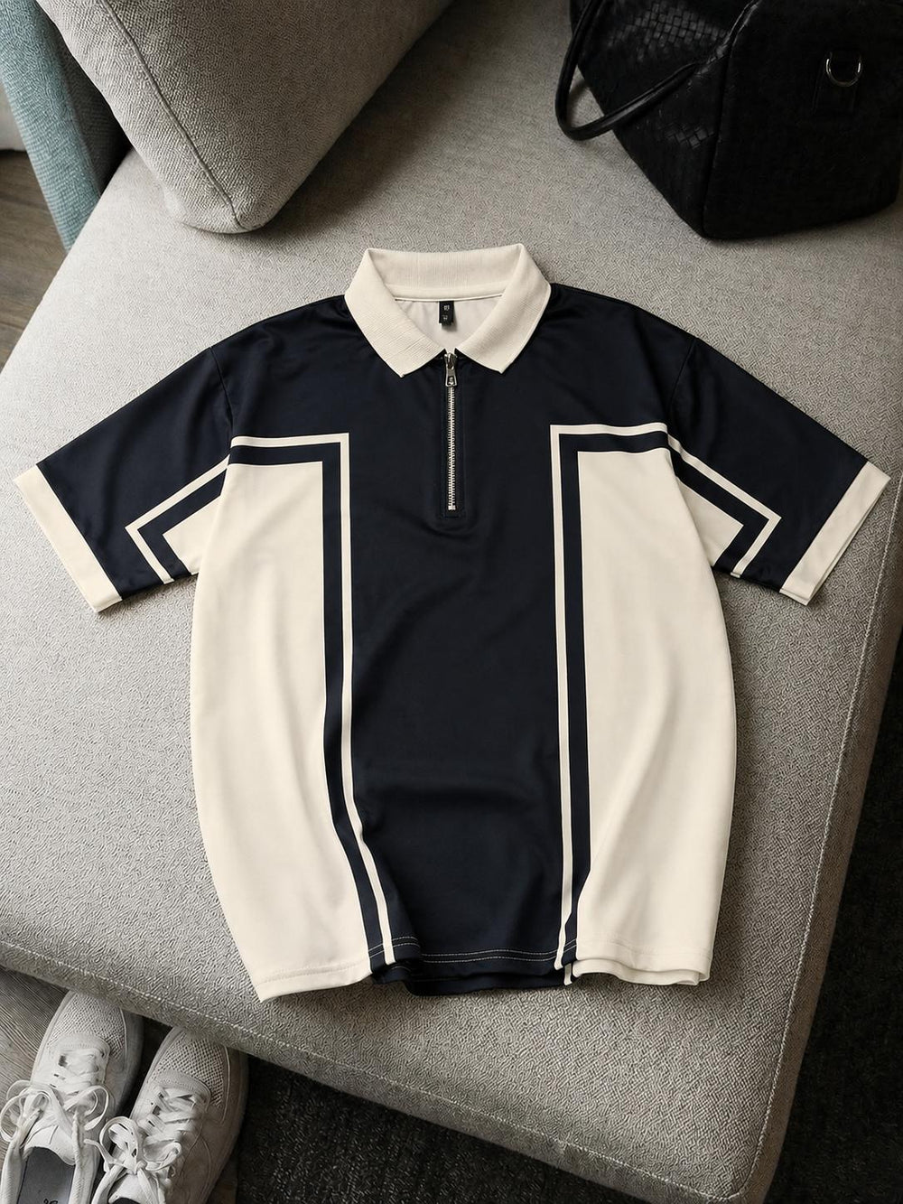 A short-sleeve polo shirt with a half-zip collar, featuring a navy blue body with cream-colored side panels and collar, laid flat on a gray upholstered bench.