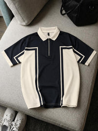 A short-sleeve polo shirt with a half-zip collar, featuring a navy blue body with cream-colored side panels and collar, laid flat on a gray upholstered bench.
