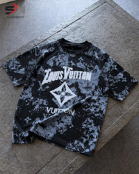 A short-sleeve t-shirt with a black and gray monogram pattern, featuring a large white graphic print on the front, laid flat on a stone surface.