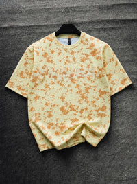 A short-sleeve crewneck t-shirt with an orange and yellow abstract paint splatter pattern, displayed on a black hanger against a dark gray textured background.