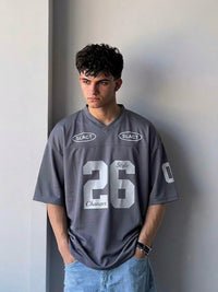 A young man wearing a gray oversized V-neck jersey with the number 26 and 'SLACT' logos, paired with light blue jeans, standing against a gray wall.