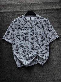 A short-sleeve crewneck t-shirt with a black and white splatter pattern, hanging on a black hanger against a dark gray textured background.