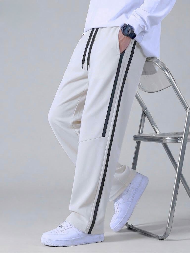 A person wearing off-white track pants with black side stripes, a white sweatshirt, and white sneakers, leaning against a metal chair in a studio setting.