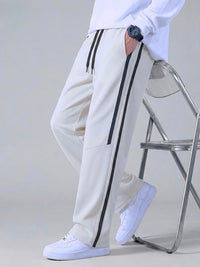 A person wearing off-white track pants with black side stripes, a white sweatshirt, and white sneakers, leaning against a metal chair in a studio setting.