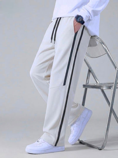 A person wearing off-white track pants with black side stripes, a white sweatshirt, and white sneakers, leaning against a metal chair in a studio setting.