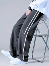 A person seated on a metal folding chair wearing dark charcoal gray wide-leg track pants with two white side stripes, a white shirt, and white sneakers against a light gray studio background.