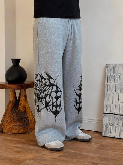 A person wearing light heather gray wide-leg sweatpants with large black abstract graphic prints on the lower legs, standing on a wood floor in an indoor room setting.