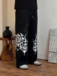 Black wide-leg sweatpants with white abstract butterfly-like graphic prints on the lower legs, shown on a model standing on a wooden floor in an indoor room setting.