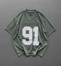 An oversized short-sleeve athletic jersey in sage green with a large white '91' printed on the front, laid flat on a dark gray background.
