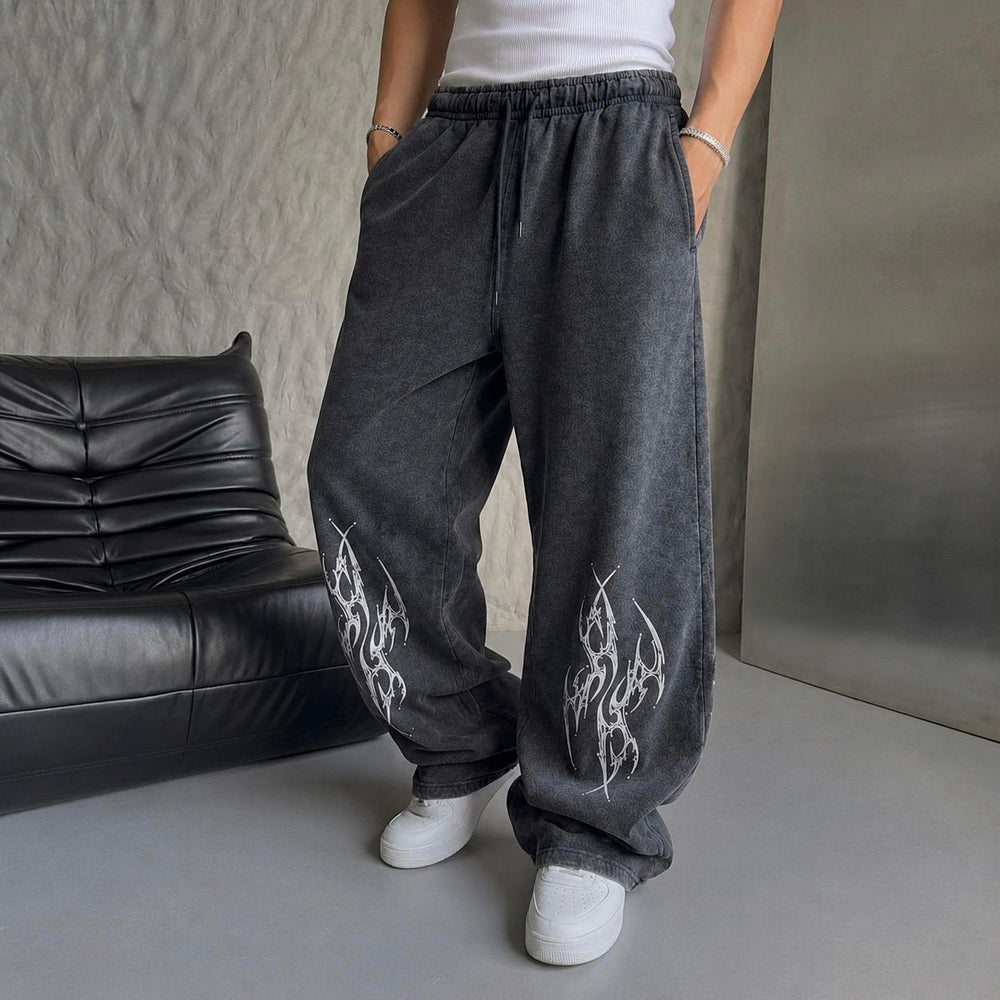 A person wearing loose, wide-leg dark gray sweatpants with white flame-style graphics on the lower legs, paired with a white top and white sneakers, standing indoors next to a black leather sofa.