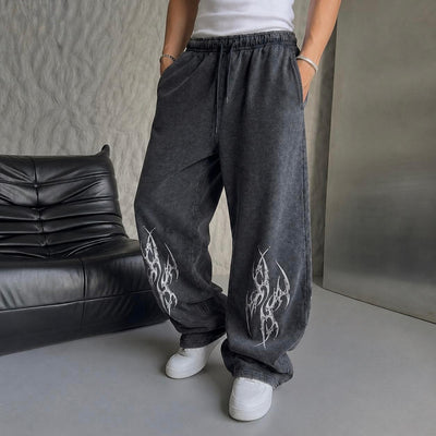 A person wearing loose, wide-leg dark gray sweatpants with white flame-style graphics on the lower legs, paired with a white top and white sneakers, standing indoors next to a black leather sofa.