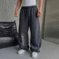 A person wearing loose, wide-leg charcoal gray sweatpants with a white script logo on the lower left leg, paired with a white t-shirt and white sneakers, standing in a studio setting with a black leather sofa.