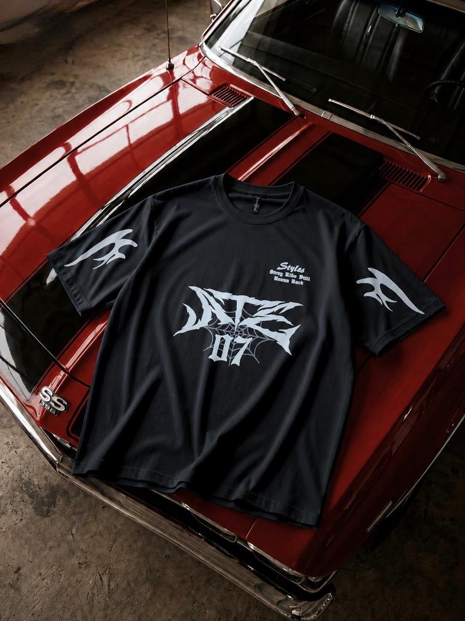 A black short-sleeve graphic t-shirt with white stylized lettering and wing motifs, laid flat on the hood of a red classic car in a garage setting.