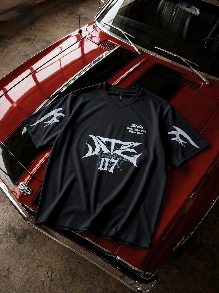 A black short-sleeve graphic t-shirt with white stylized lettering and wing motifs, laid flat on the hood of a red classic car in a garage setting.