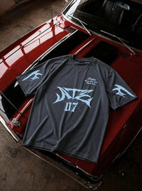 A dark gray graphic t-shirt with light blue designs, featuring a large stylized 'NZE 07' on the front and bird graphics on the sleeves, laid flat on the hood of a red classic car.