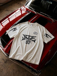 An off-white short-sleeve graphic t-shirt with black designs, displayed on the hood of a red classic car in a garage setting.