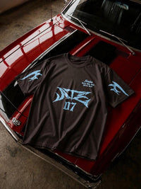 A dark brown t-shirt with a light blue graphic print, laid flat on the hood of a red classic car.
