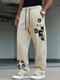 A person wearing beige drawstring sweatpants with black floral prints and the word 'Chicago' on the thigh, paired with white sneakers in a concrete setting.