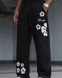 Black wide-leg sweatpants with white floral prints and the word 'Chicago' on the thigh, shown on a model in a gray indoor setting.