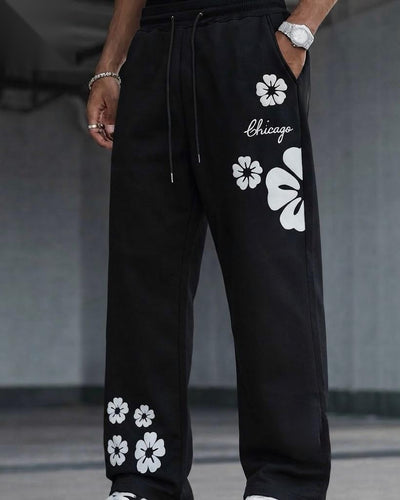 Black wide-leg sweatpants with white floral prints and the word 'Chicago' on the thigh, shown on a model in a gray indoor setting.