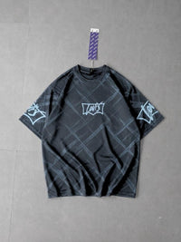 A dark gray short-sleeve t-shirt with a subtle diagonal grid pattern, featuring a white graphic logo on the chest and sleeve prints on both sleeves. The shirt is displayed flat against a gray tiled background with a purple patterned hangtag attached to the collar.
