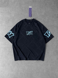 A black short-sleeve t-shirt with light blue graphic prints on the chest, sleeves, and collar, hanging on a purple patterned tag against a gray tiled floor.