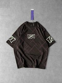 A dark brown short-sleeve t-shirt with a subtle plaid pattern, featuring a white graphic logo on the chest and sleeve, laid flat on a gray background with a hanging tag.