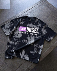 A Diesel Denim Division t-shirt with a black and gray distressed paint-splatter pattern, featuring a purple and white logo on the chest, laid flat on a gray stone surface.