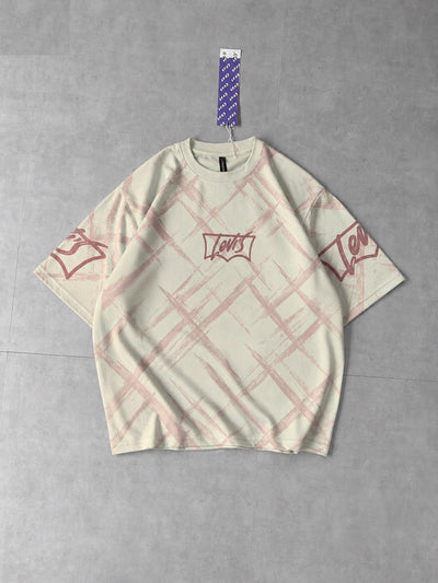 A cream-colored Levi's short-sleeve t-shirt with a pink plaid pattern and red graphic prints on the chest and sleeves, laid flat on a gray concrete floor.
