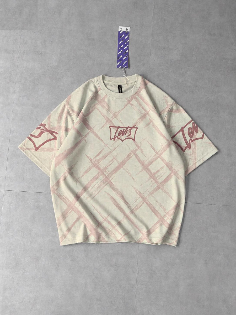 A cream-colored Levi's short-sleeve t-shirt with a pink plaid pattern and red graphic prints on the chest and sleeves, laid flat on a gray concrete floor.