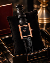 A Cartier watch with a rectangular rose gold case, black dial, and black alligator leather strap, displayed on a black velvet cushion inside an open black and gold Cartier box.