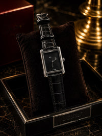 A luxury rectangular analog watch with a black dial and black crocodile-embossed strap, displayed on a black velvet cushion inside an open black presentation box.