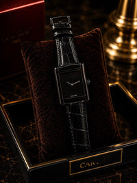 A black rectangular Cartier watch with a black alligator strap, displayed on a dark brown velvet cushion inside a black and gold Cartier presentation box.