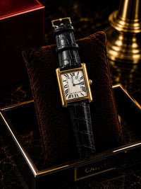 A Cartier Tank watch with a gold-tone case, white dial, and black crocodile-embossed strap, displayed on a brown velvet cushion inside an open black and gold Cartier box.