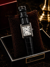 A luxury watch with a silver-toned rectangular case, white dial with black Roman numerals, and a black alligator leather strap, displayed on a dark brown velvet cushion inside a black presentation box.