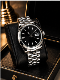 A stainless steel wristwatch with a black dial, black date window, and silver link bracelet, displayed on a black leather watch cushion inside a black presentation box.