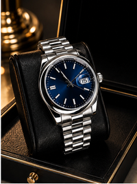 A stainless steel wristwatch with a deep blue dial and date window, resting on a black leather cushion inside a luxury watch box with gold trim.